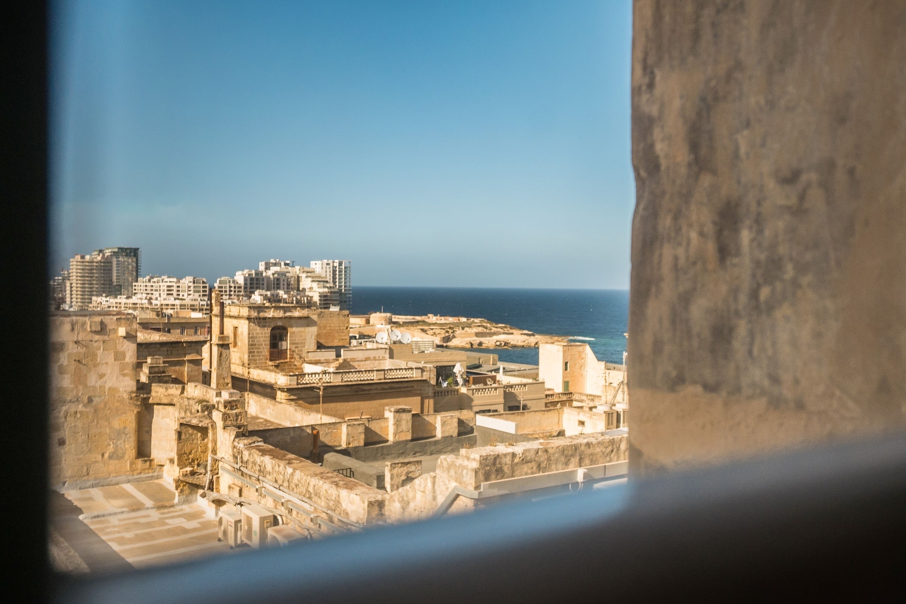 Une vue sur un paysage urbain côtier, prise depuis une fenêtre, qui présente un mélange de bâtiments historiques et modernes, un ciel dégagé et la mer en arrière-plan.