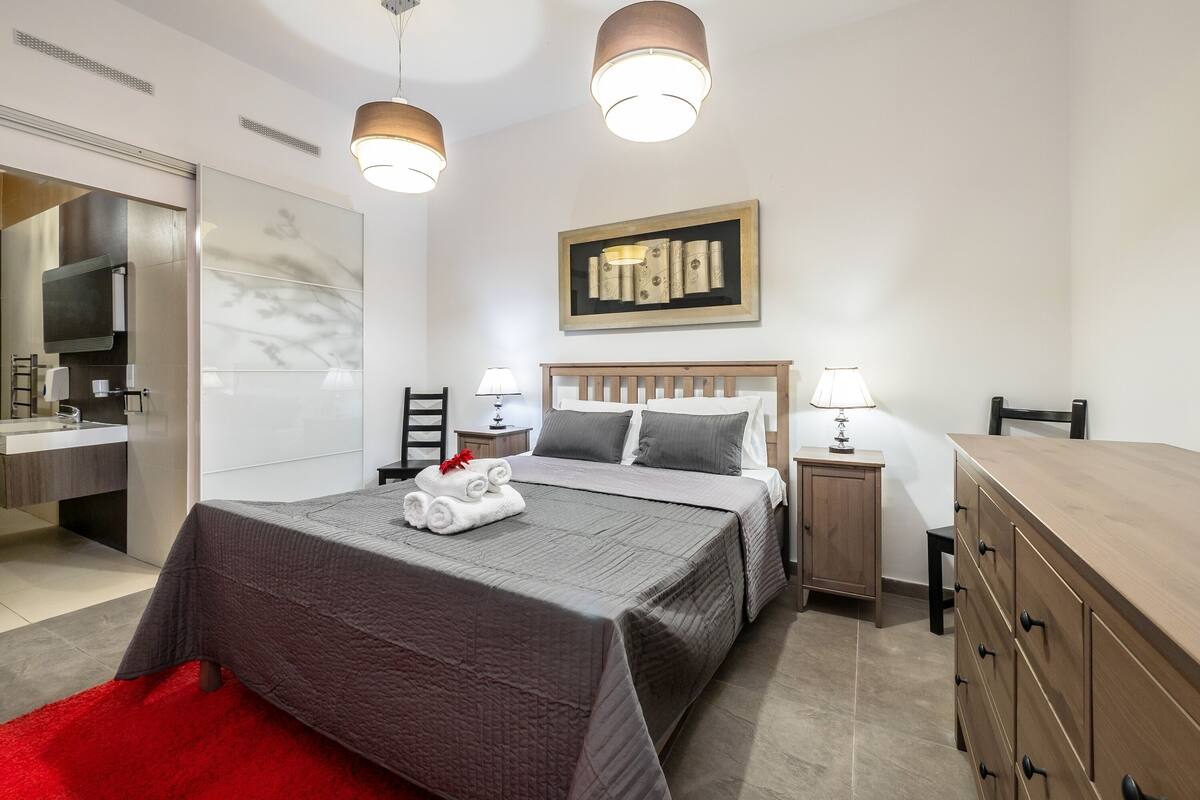 A modern hotel room with a large bed, bedside tables, a wardrobe, and a bathroom with a mirror and sink.