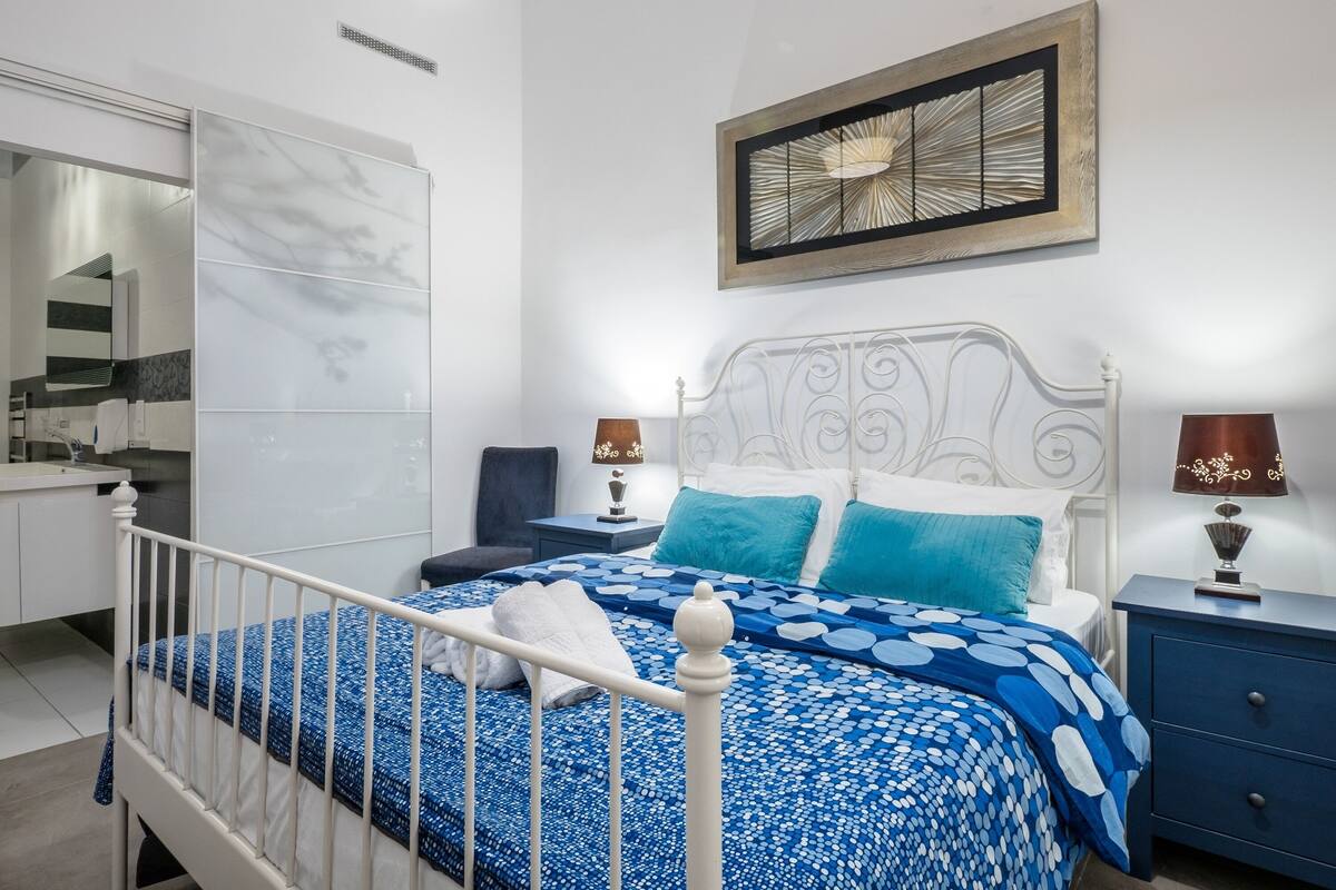 A bedroom with a white metal bed frame, a blue patterned bedspread, two bedside lamps, a nightstand, and a wall-mounted picture.