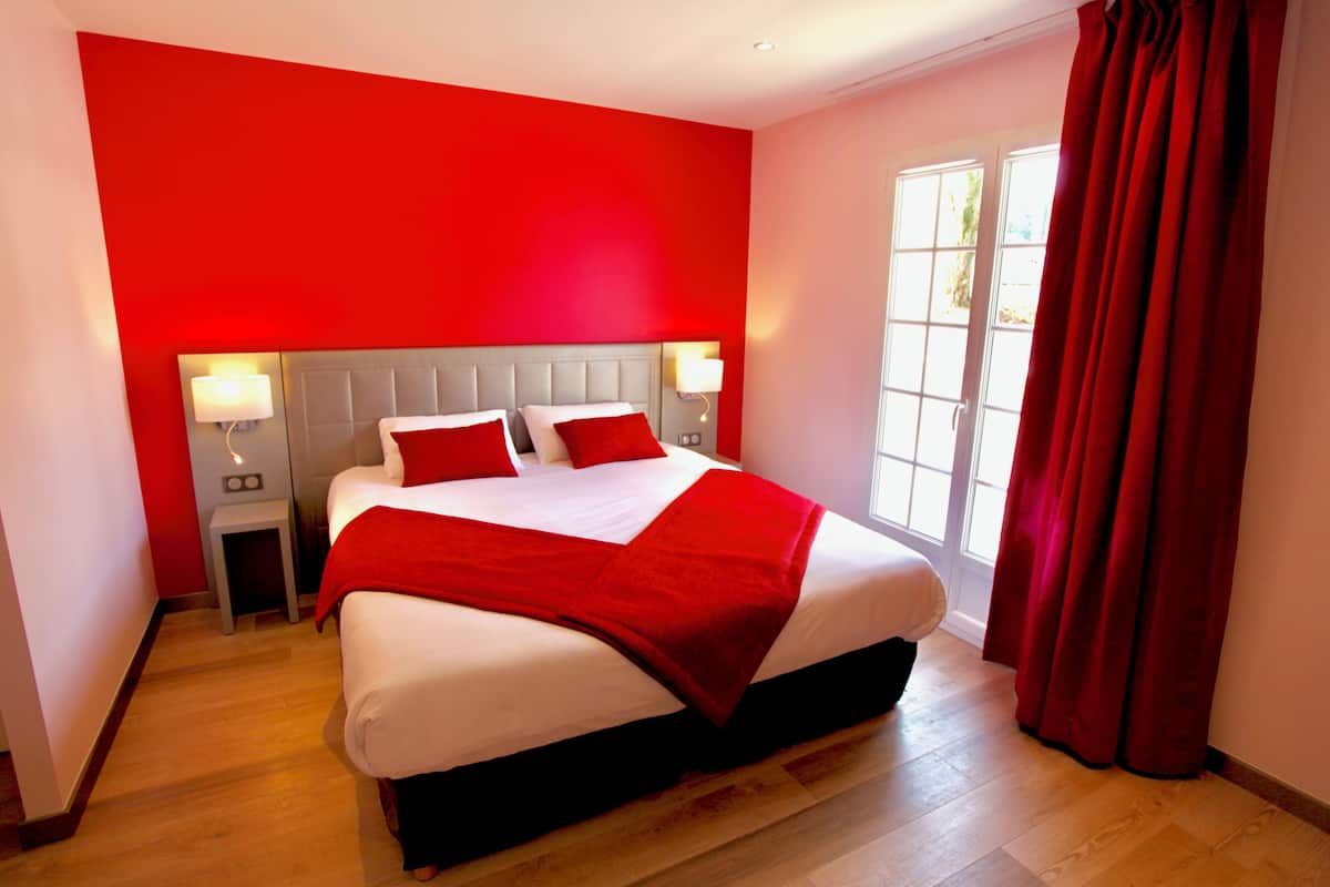 Una habitación de hotel con una cama grande, dos mesitas de noche, una pared de acento roja y una ventana con cortinas.