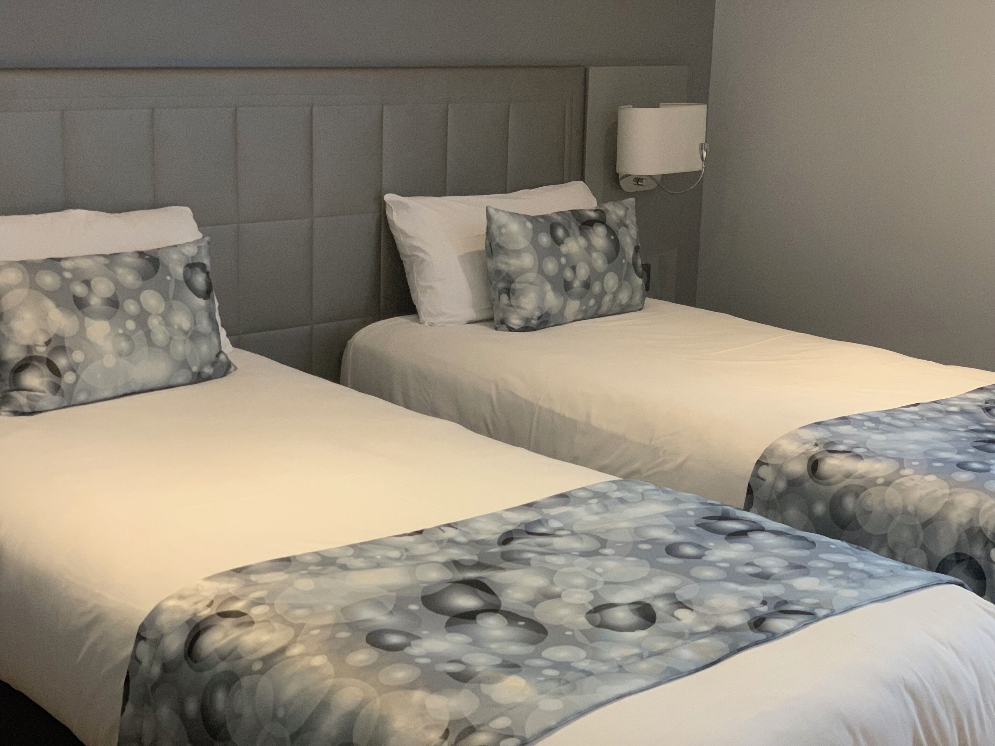 A hotel room with two beds, grey headboard, white bedding, and a wall-mounted lamp.