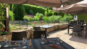 Un patio al aire libre con mesas y sillas, un muro de piedra y un jardín con diversas plantas y flores.