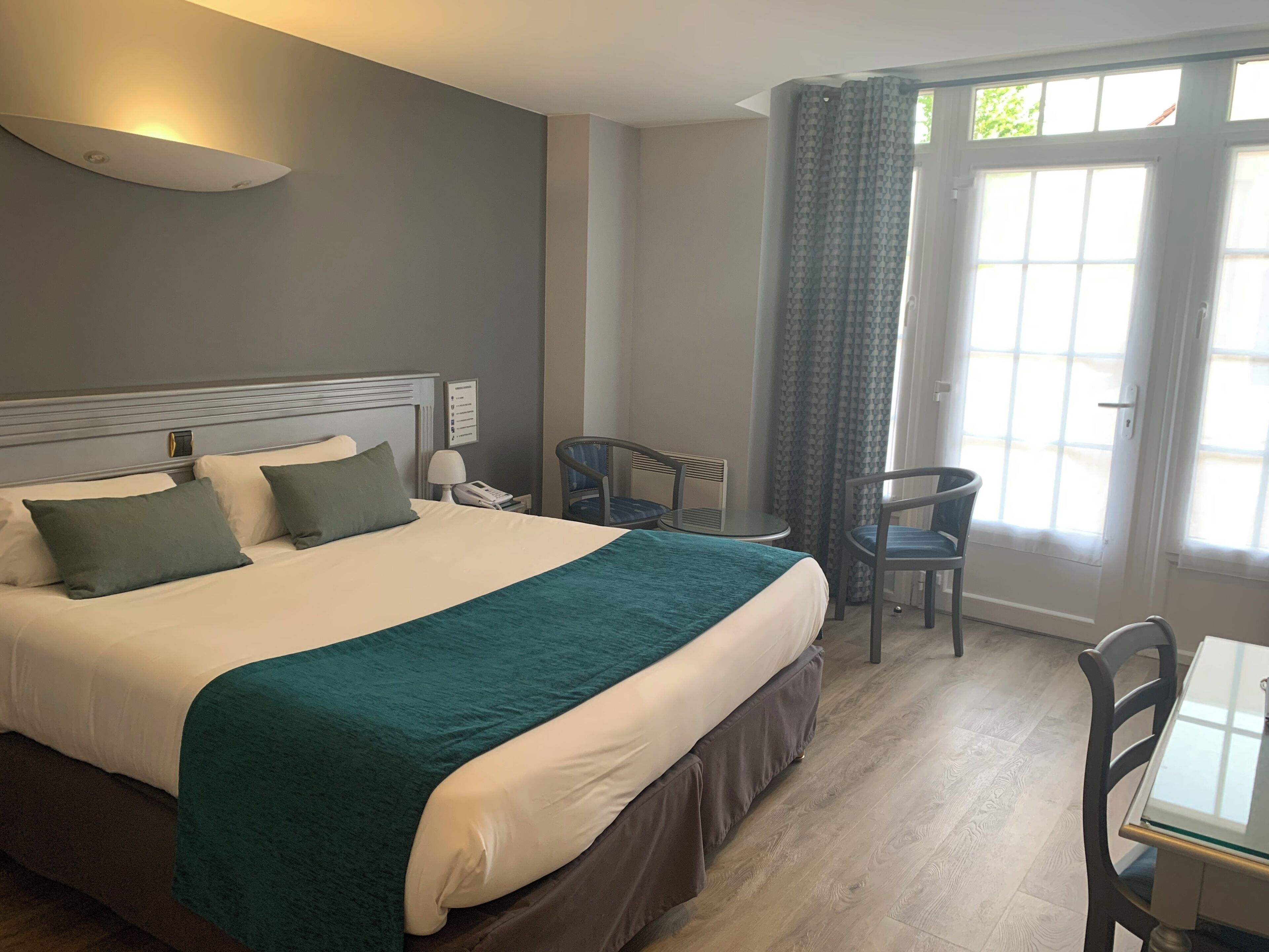 A hotel room with a bed, bedside table, lamp, two chairs, and a window with curtains.