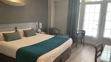 A hotel room with a bed, bedside table, lamp, two chairs, and a window with curtains.