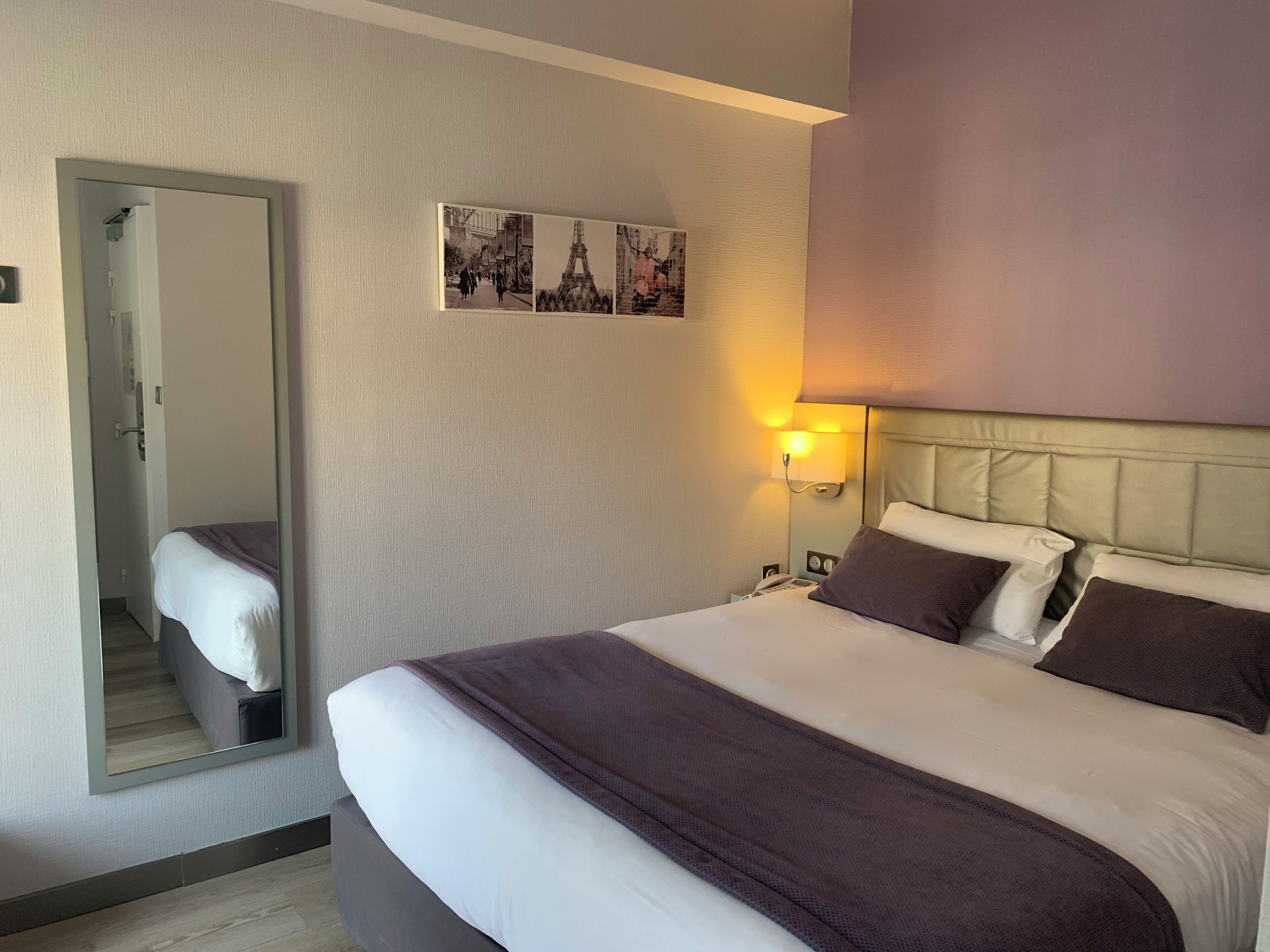 A hotel room with a large bed, a bedside lamp, a mirror, and a wall-mounted picture of Paris.