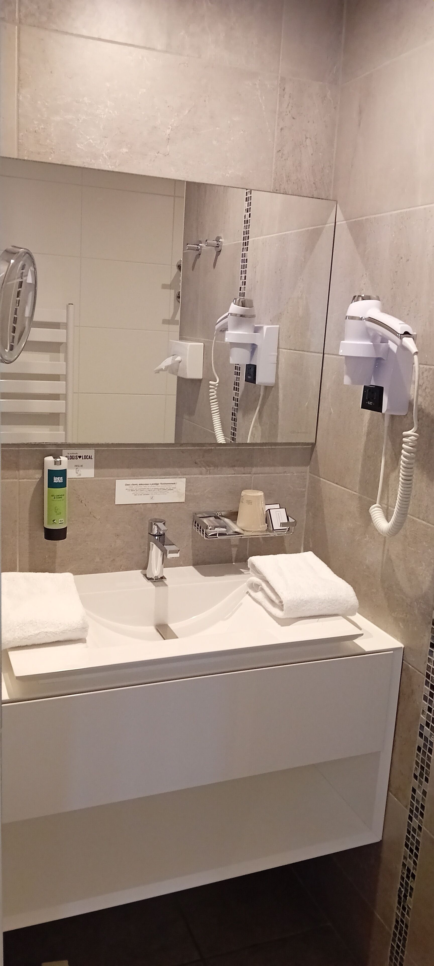 Un baño moderno con un espejo grande, un lavabo, un perchero para toallas, un secador de pelo y una botella de loción.