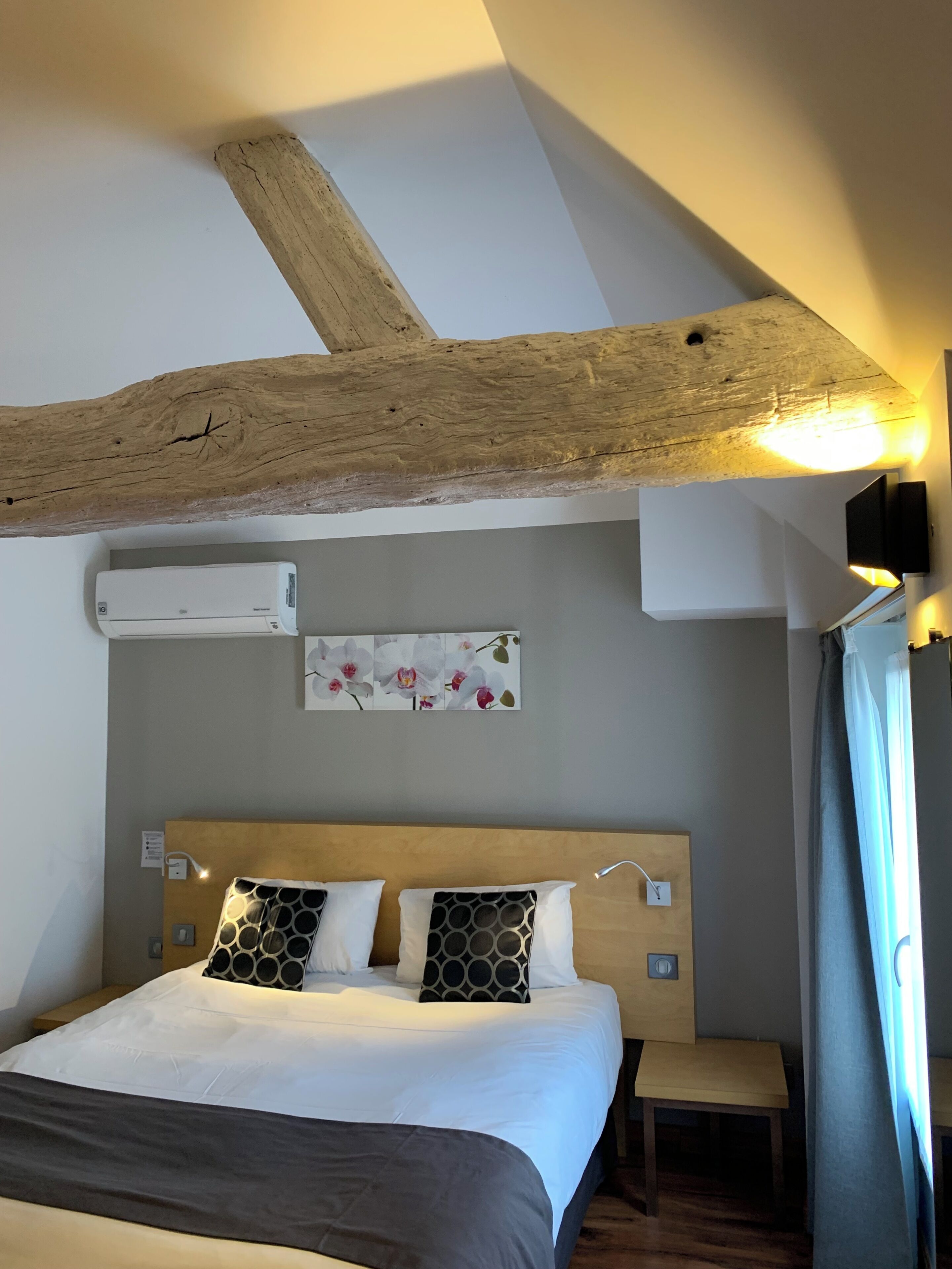 A hotel room with a wooden ceiling beam, a bed with patterned pillows, a small bedside table, and a wall-mounted air conditioner.