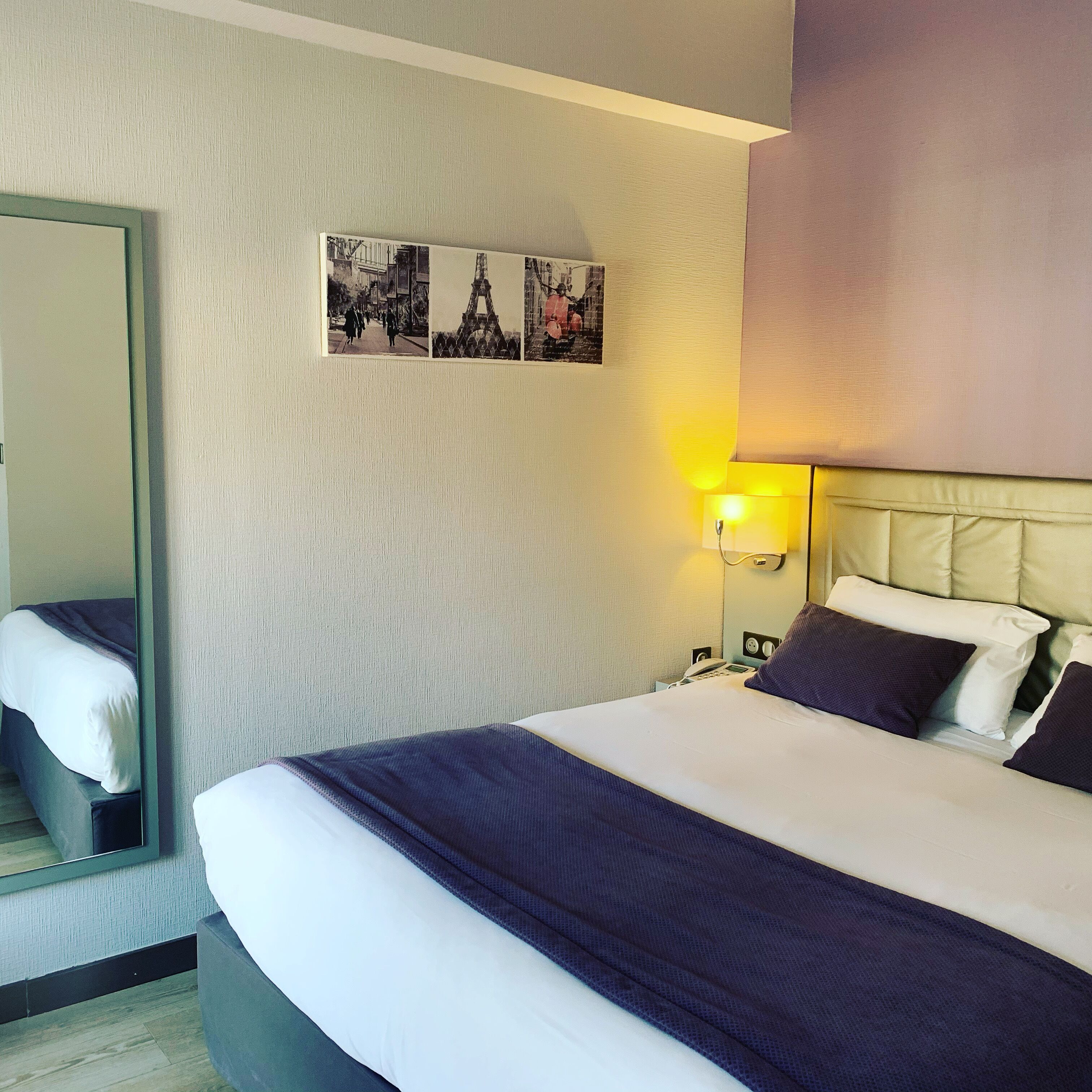 Une chambre d’hôtel avec un lit, une table de chevet, une lampe, un miroir et un mur orné de trois tableaux.
