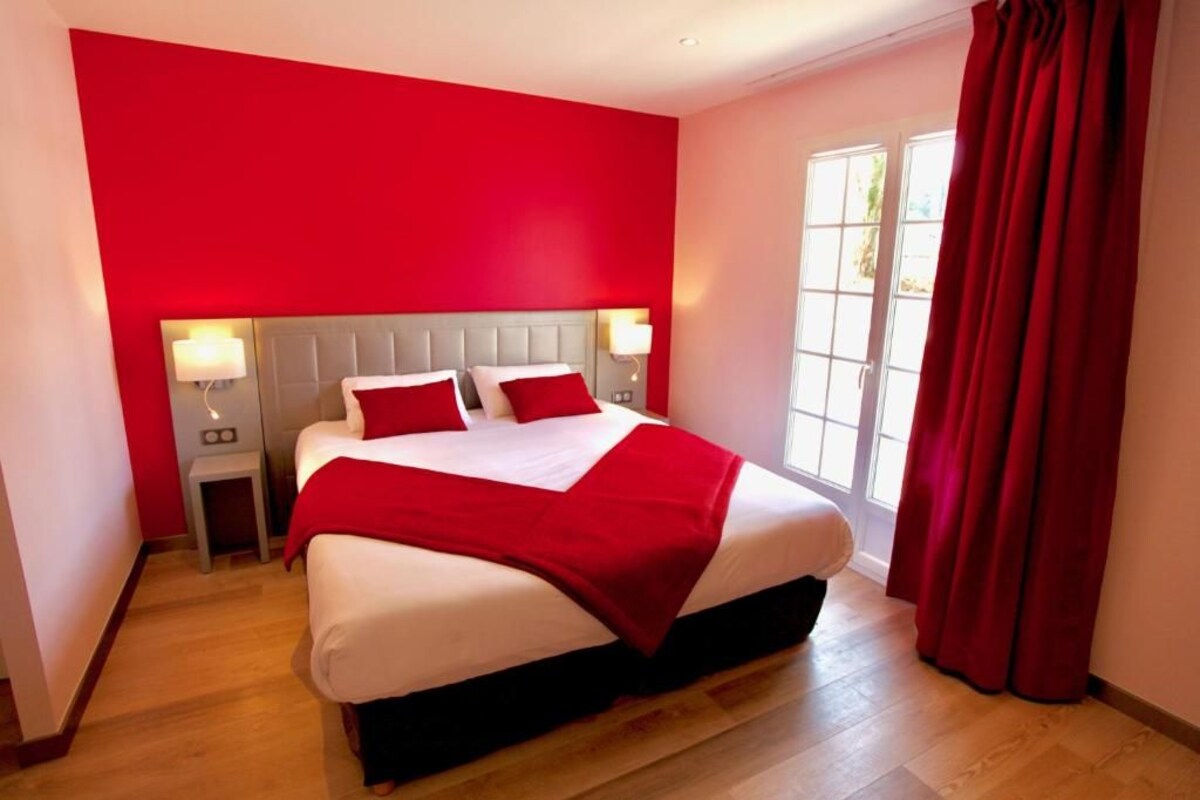 Habitación de hotel con una cama grande, dos mesitas de noche con lámparas, una pared de acento roja y un ventanal con cortinas.