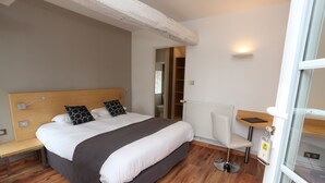 Un dormitorio con cabecera de madera, cama con ropa de cama blanca y oscura, mesita de noche con lámpara, una silla y vistas al baño.