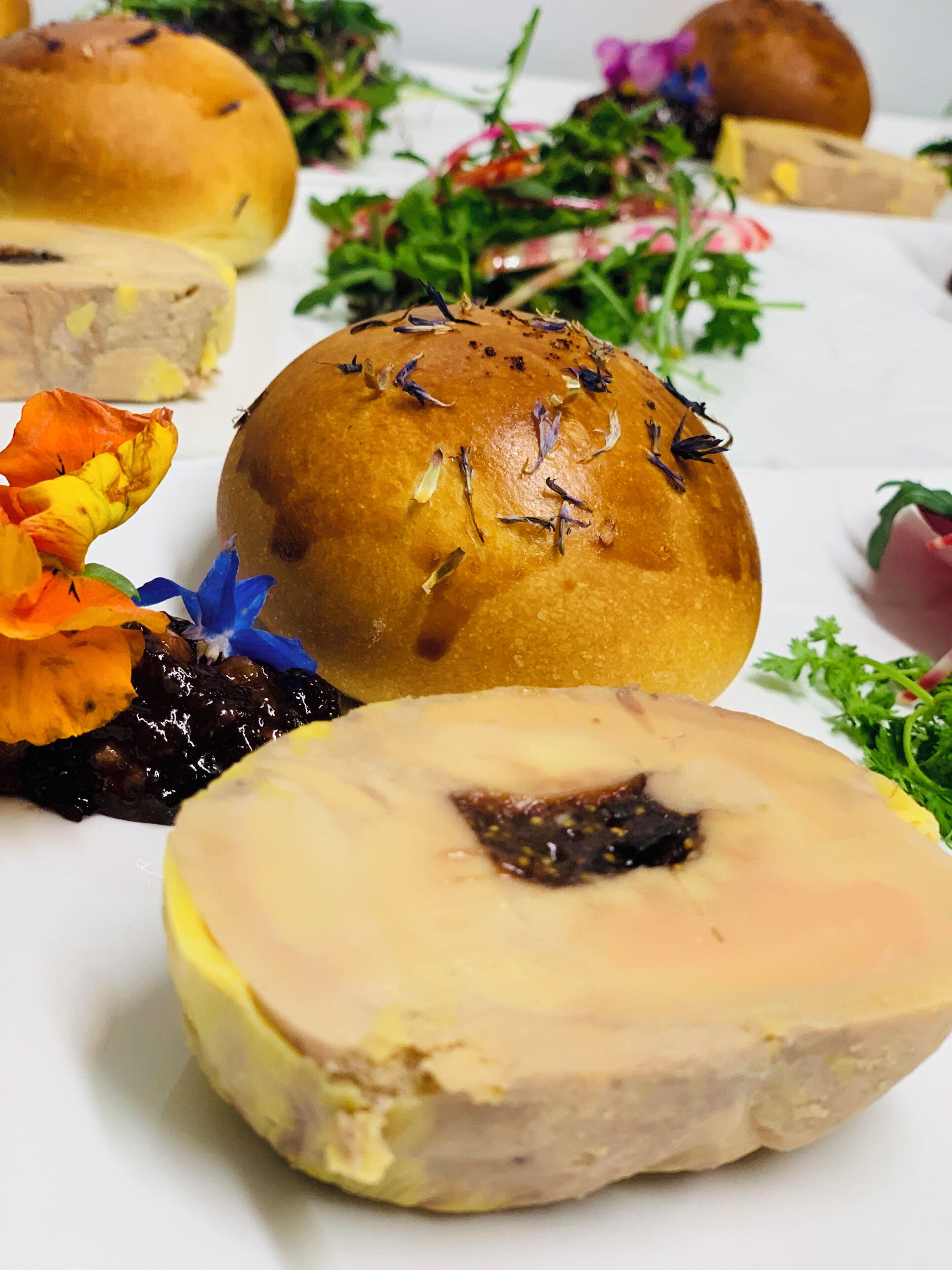 A variety of food items including a bread roll, a sliced meat dish, and some flowers.