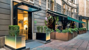 Entrada de hotel con puerta de vidrio, zona de estar exterior con macetas y un cartel con el nombre del hotel.