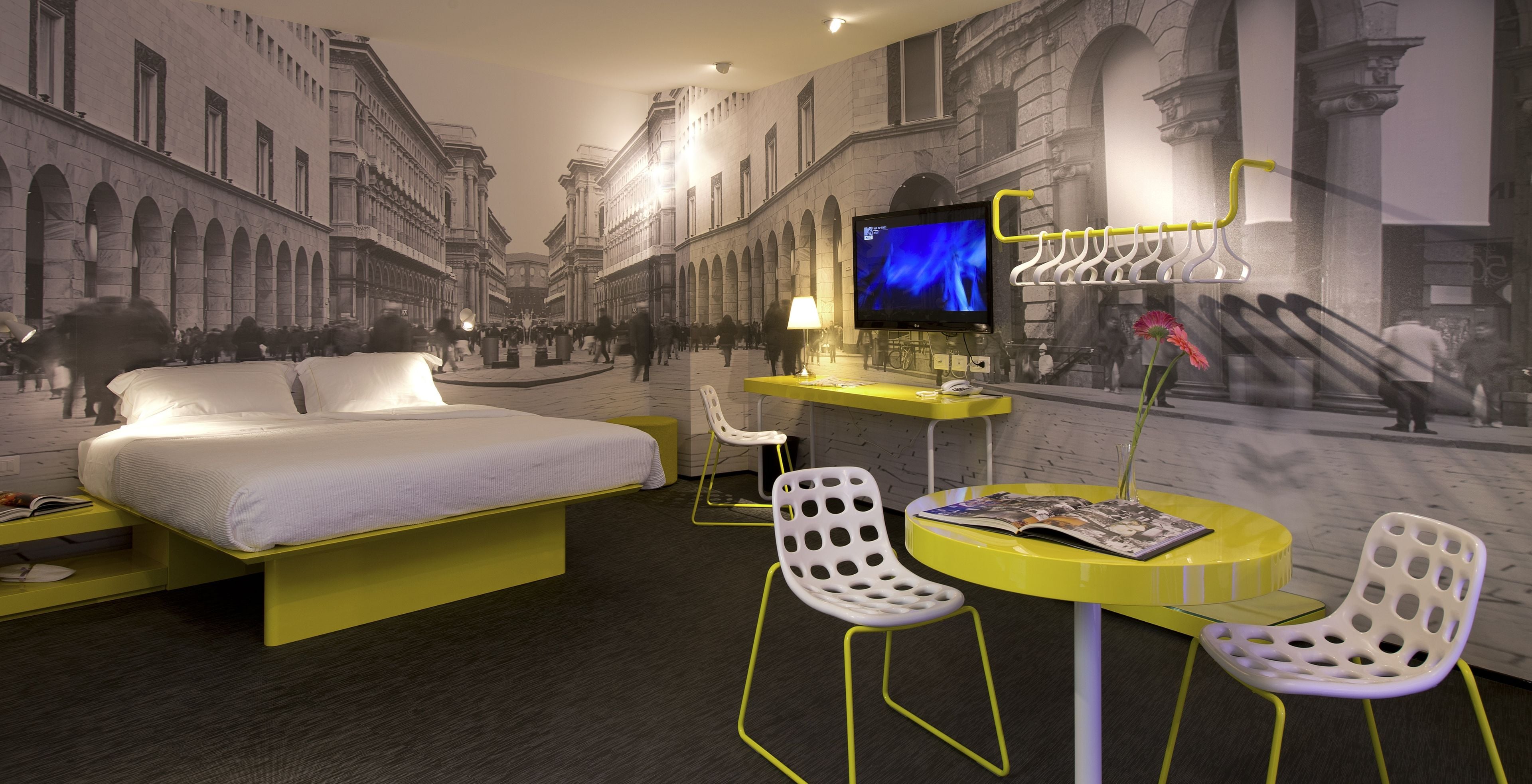 Habitación de hotel con cama, escritorio, dos sillas, televisor y un mural con una escena callejera.
