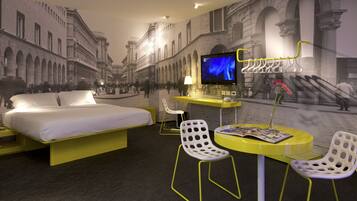 Habitación de hotel con cama, escritorio, dos sillas, televisor y un mural con una escena callejera.