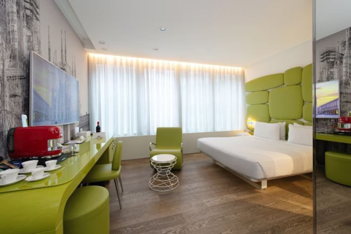 Une chambre d’hôtel avec un lit, un bureau doté d’une machine à café, une chaise et une grande fenêtre avec des rideaux.