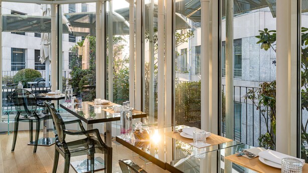 Un restaurante moderno con mesas y sillas de vidrio, ventanas amplias y plantas de interior.