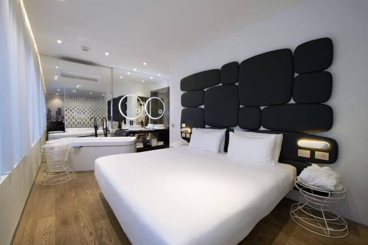 Una habitación de hotel moderna con una cama grande, una mesita de noche, un espejo y un baño visible al fondo.