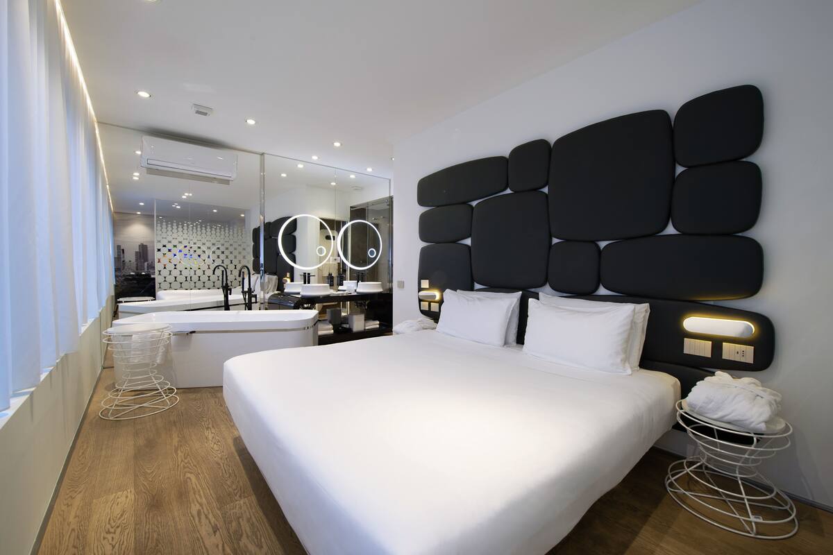 Habitación de hotel moderna con una cama grande, una mesita de noche, un espejo y un baño al fondo.