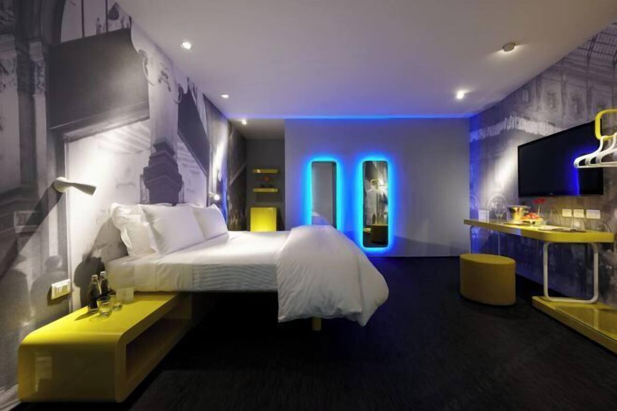 Una habitación de hotel moderna con cama, mesita de noche, lámpara y televisor sobre un mueble.