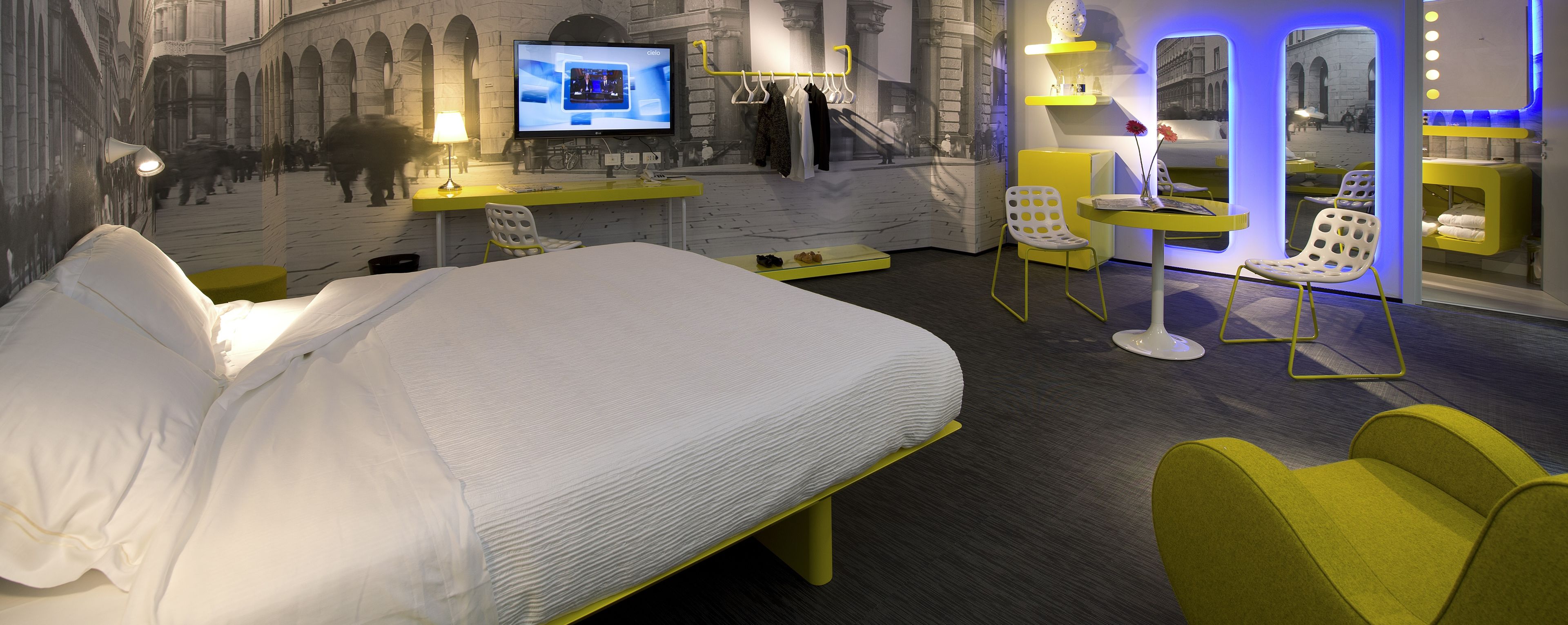 Une chambre d’hôtel comprenant un lit, un bureau avec un ordinateur, un coin salon avec des fauteuils et un mur orné d’une fresque.