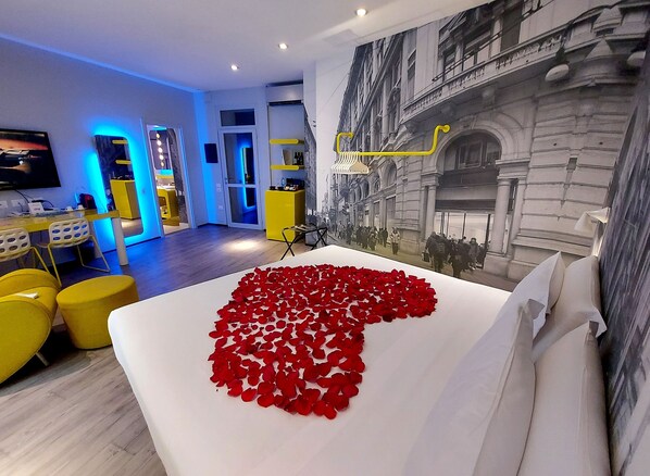 Une chambre d’hôtel avec un lit parsemé de pétales de roses rouges, un coin repas moderne aux couleurs jaunes et une grande fresque représentant un paysage urbain.