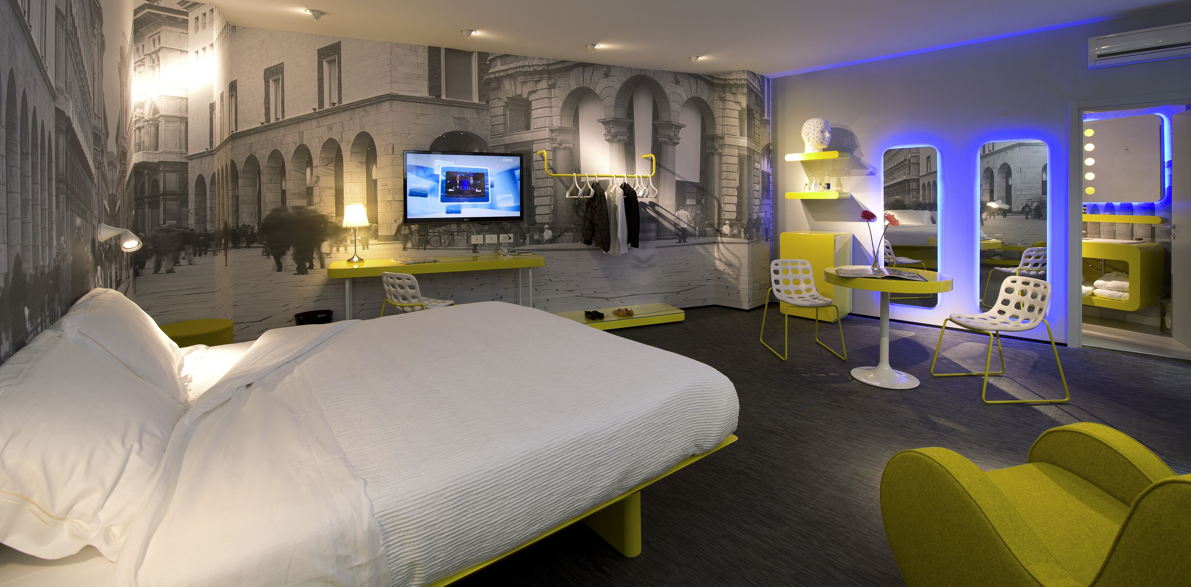 A modern hotel room with a large bed, a desk with a computer, a chair, and a mirror.