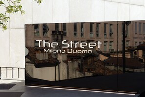 Une façade d’immeuble sur laquelle on peut lire « The Street Milano Duomo » et qui reflète une scène de rue.