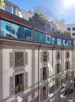Un edificio de varios pisos con amplios ventanales, balcones con plantas y una estructura moderna de vidrio en el piso superior.