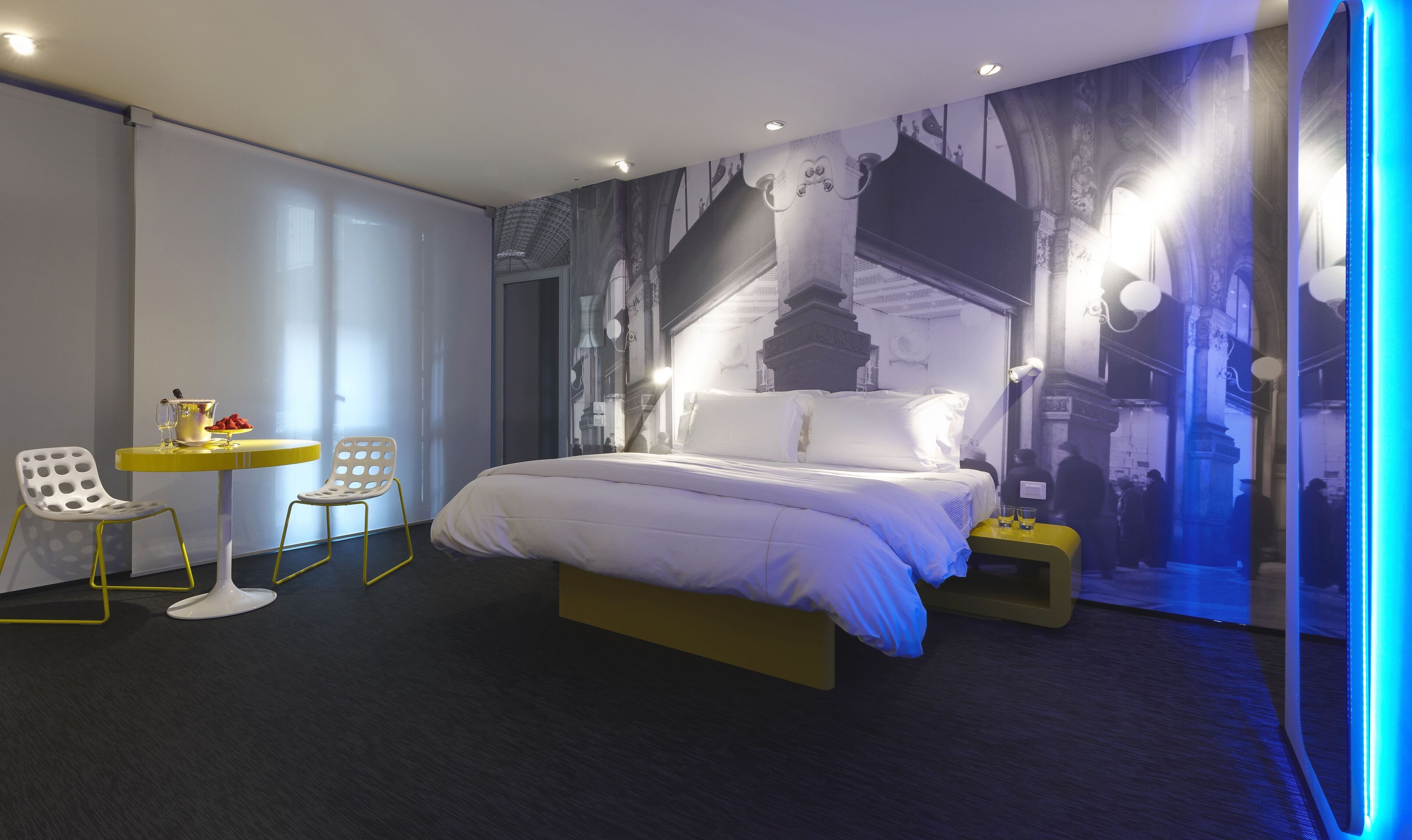 Una habitación de hotel moderna con una cama grande, una mesita redonda pequeña con dos sillas y un mural en la pared.
