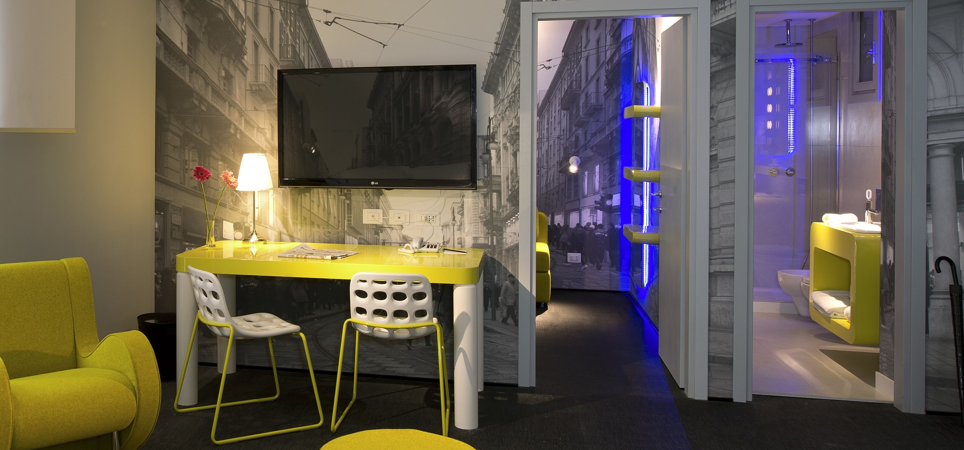 A modern hotel room with a yellow desk, two white chairs, a flat-screen TV, and a bathroom with a blue-lit shower.