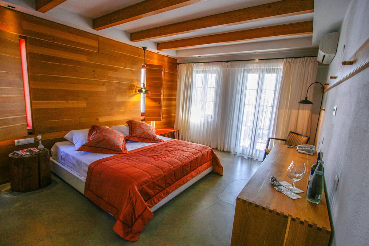 A hotel room with a wooden ceiling, a bed with orange bedding, a wooden desk with a bottle and glass, and a window with curtains.