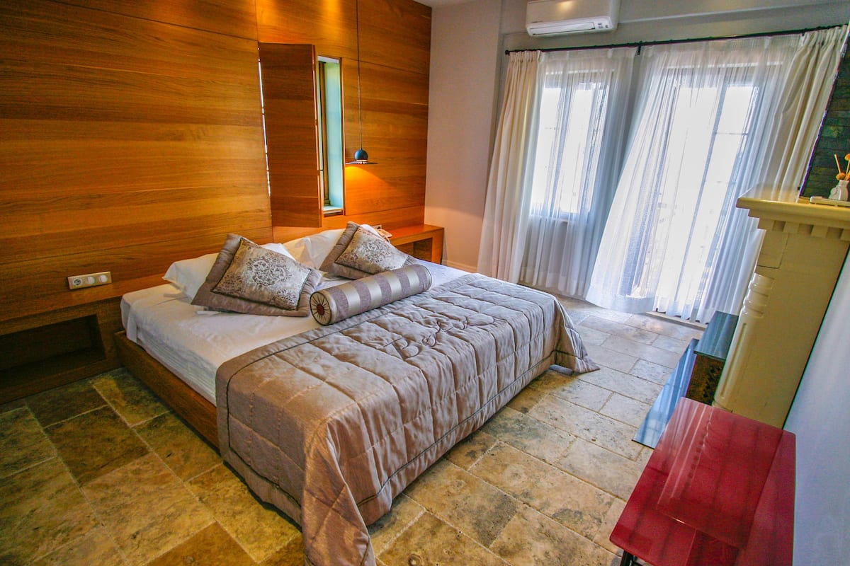 A bedroom with a wooden headboard, a large bed with multiple pillows, a bedside table, and a window with curtains.
