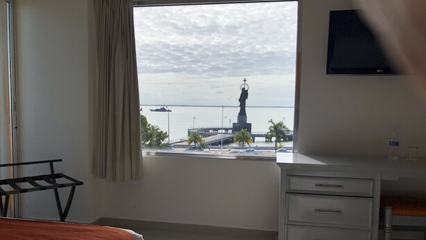 Habitación de hotel con un ventanal grande que ofrece vistas a una estatua y un barco. Hay una cama, un escritorio y un televisor montado en la pared.
