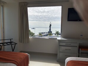 A hotel room with a large window offering a view of a statue and a ship. There is a bed, a desk, and a TV mounted on the wall. - Hotel del Parque (Ciudad del Carmen)