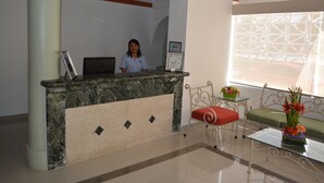 Mostrador de recepción con una recepcionista, una computadora portátil y un certificado enmarcado. Una mesa de centro de vidrio con un jarrón de flores y un sofá verde con un cojín rojo.