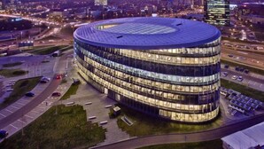 Un edificio moderno y circular con fachada de vidrio y techo plano, rodeado de una zona de estacionamiento y calles.