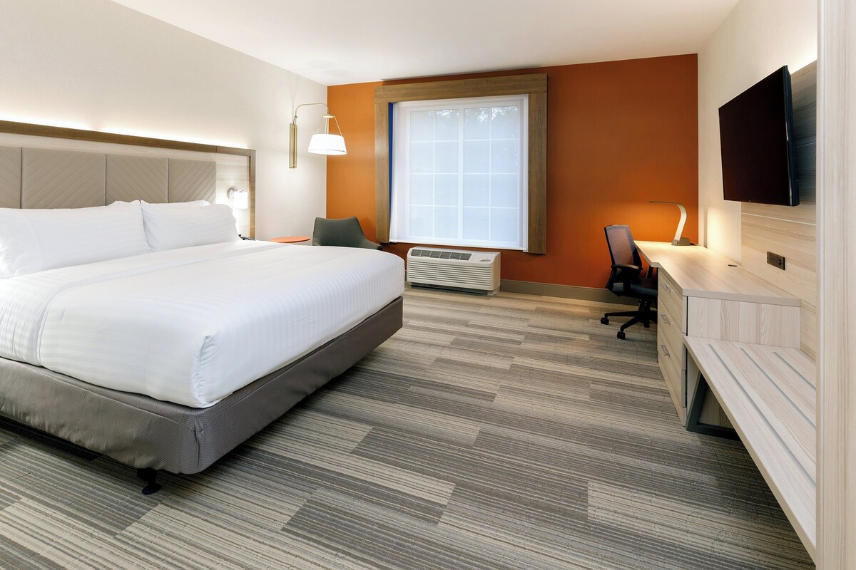 A hotel room with a large bed, a desk with a chair, a flat-screen TV, and a window with blinds.