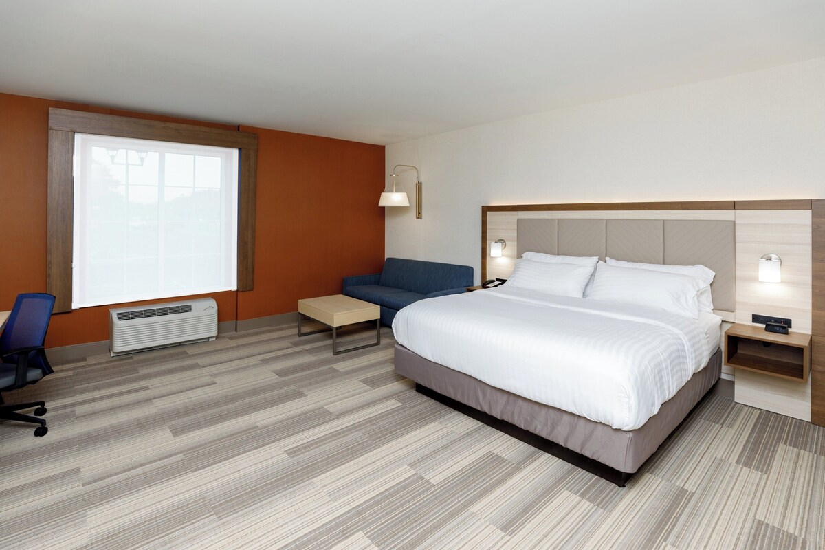 A hotel room with a large bed, a desk, a chair, a standing lamp, and a window.