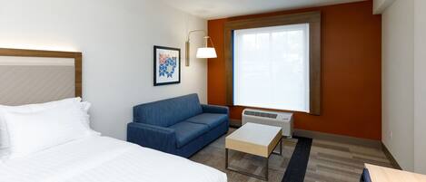 A hotel room with a bed, a blue sofa, a small table, a wall-mounted lamp, and a large window.