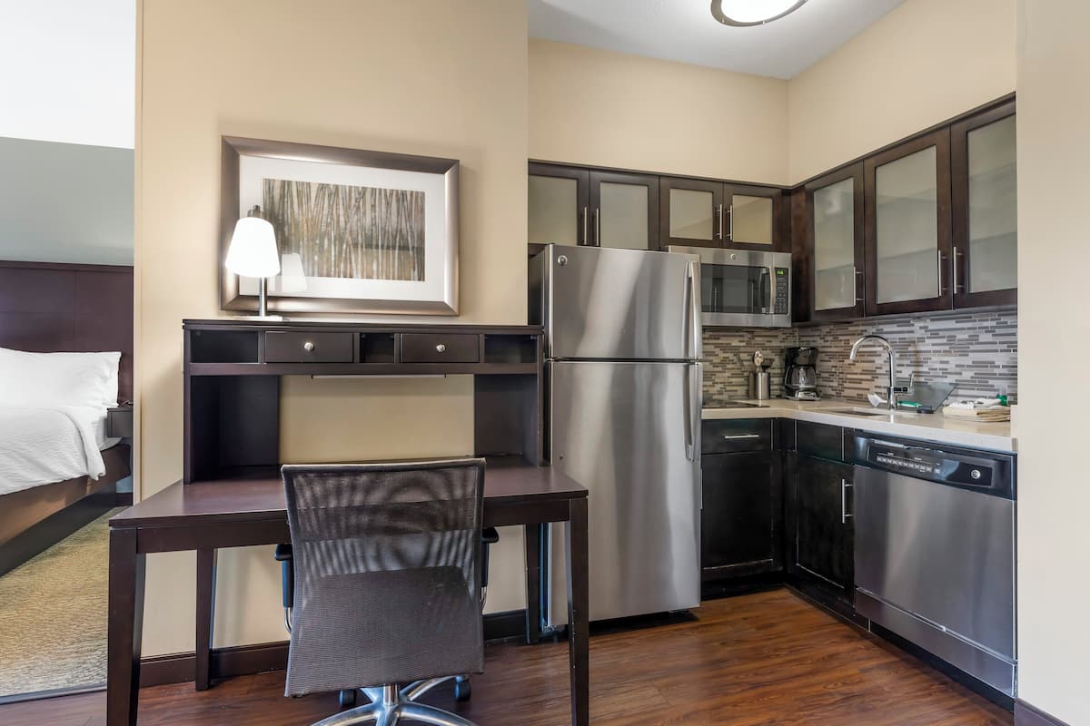 A compact hotel room with a kitchenette, a bed with white linens, a desk with a chair, and a framed picture on the wall.