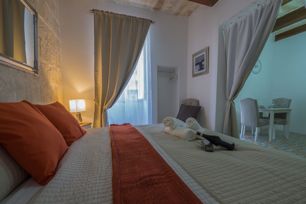 A hotel room with a bed, a desk, a chair, and a bathroom visible through a window.