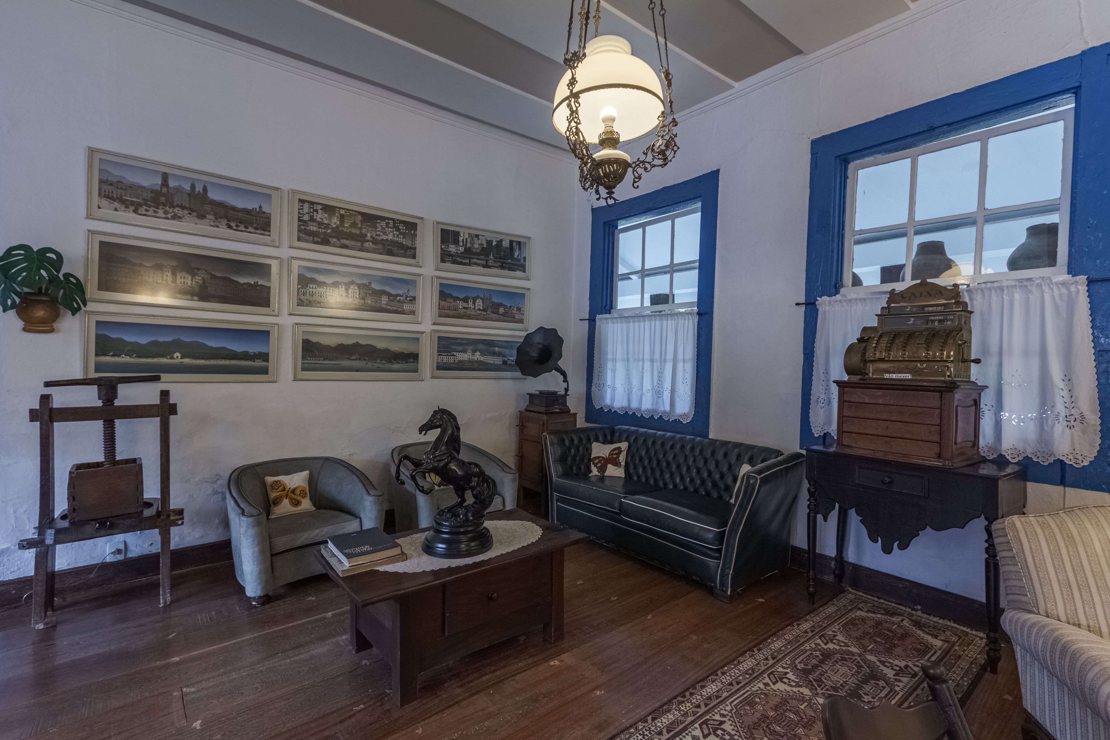 A room with a wooden floor, a blue-framed window, a chandelier, a sofa, a small table, and several framed pictures on the wall.