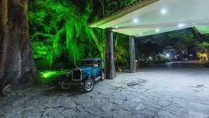 Ein Oldtimer steht unter einer Überdachung auf einem gepflasterten Platz, umgeben von üppigem Grün und beleuchtet von grünen Lichtern.