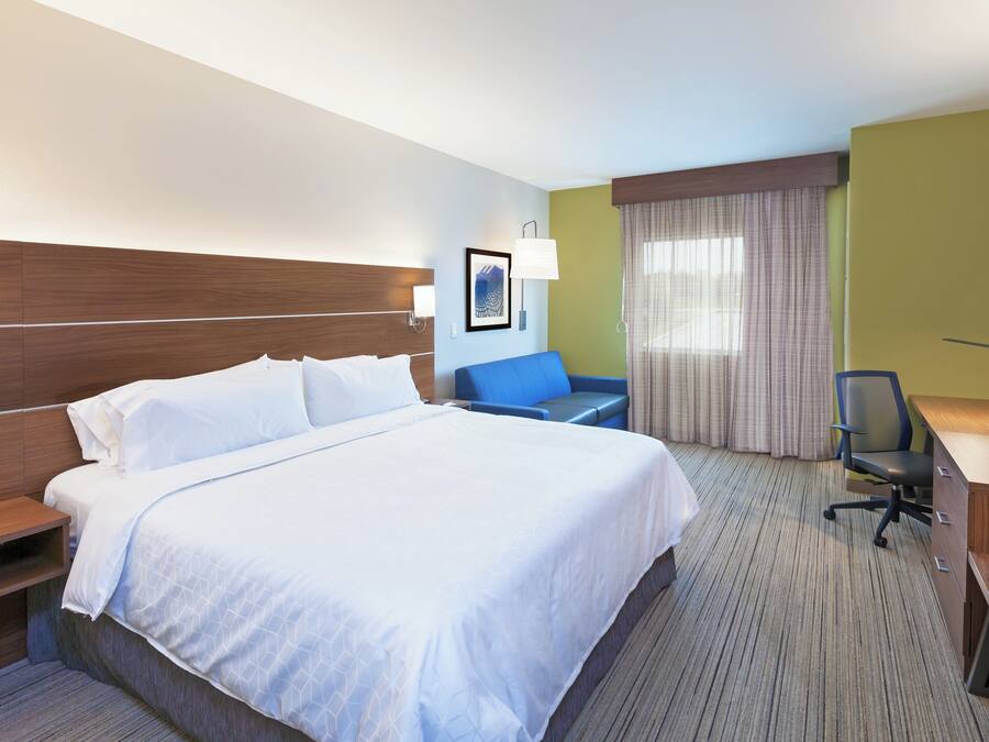 Habitación de hotel con una cama grande, un escritorio con computadora, una silla, una lámpara y un ventanal con cortinas.