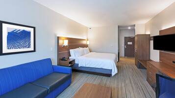 A hotel room with a bed, a blue sofa, a wooden desk, a television, and a framed artwork on the wall.