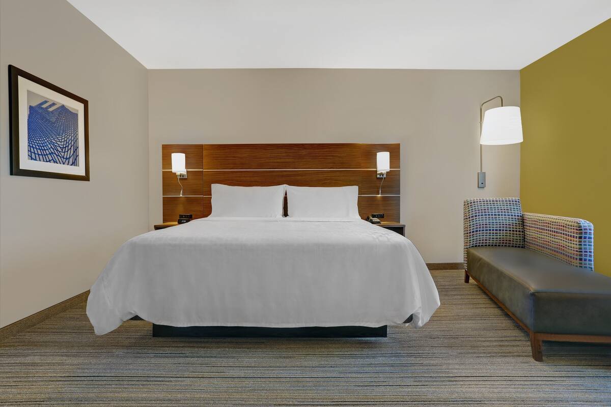 A hotel room with a large bed, a headboard with integrated lighting, a bedside table, a lamp, a sofa, and a framed artwork on the wall.
