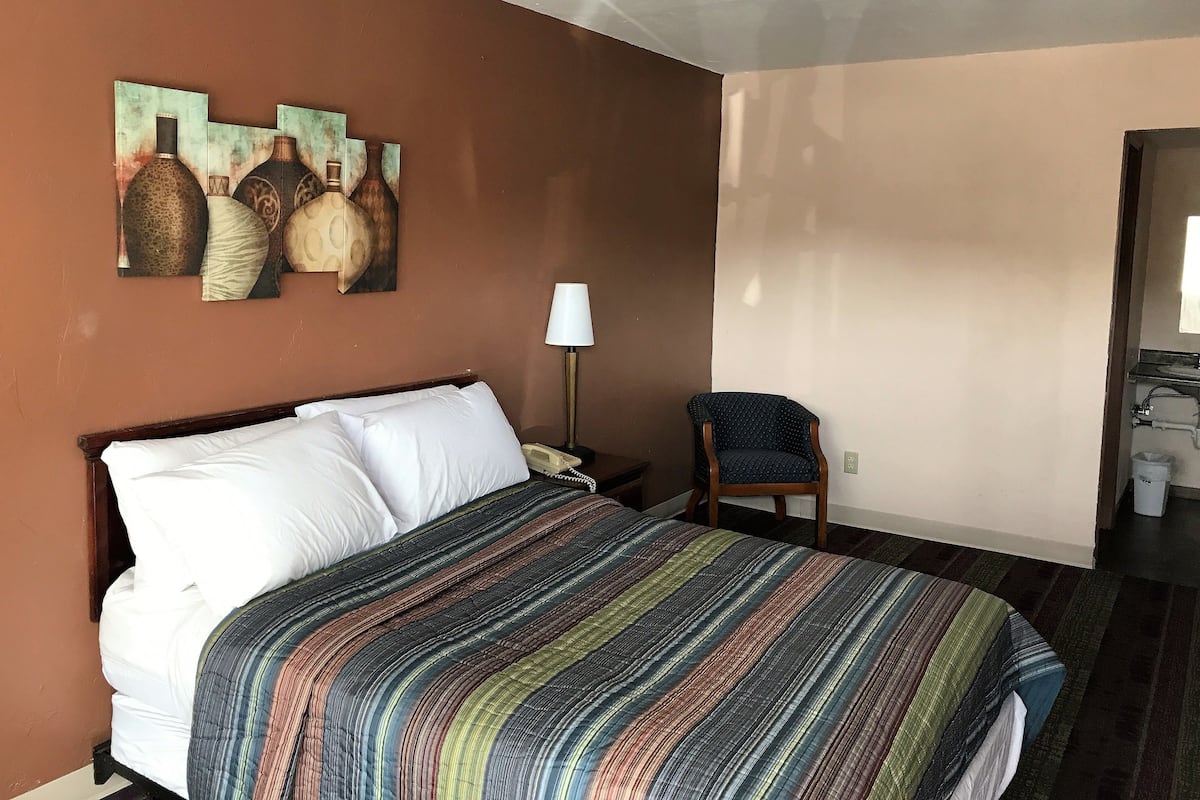 A hotel room with a bed, a chair, a lamp, and artwork on the wall.