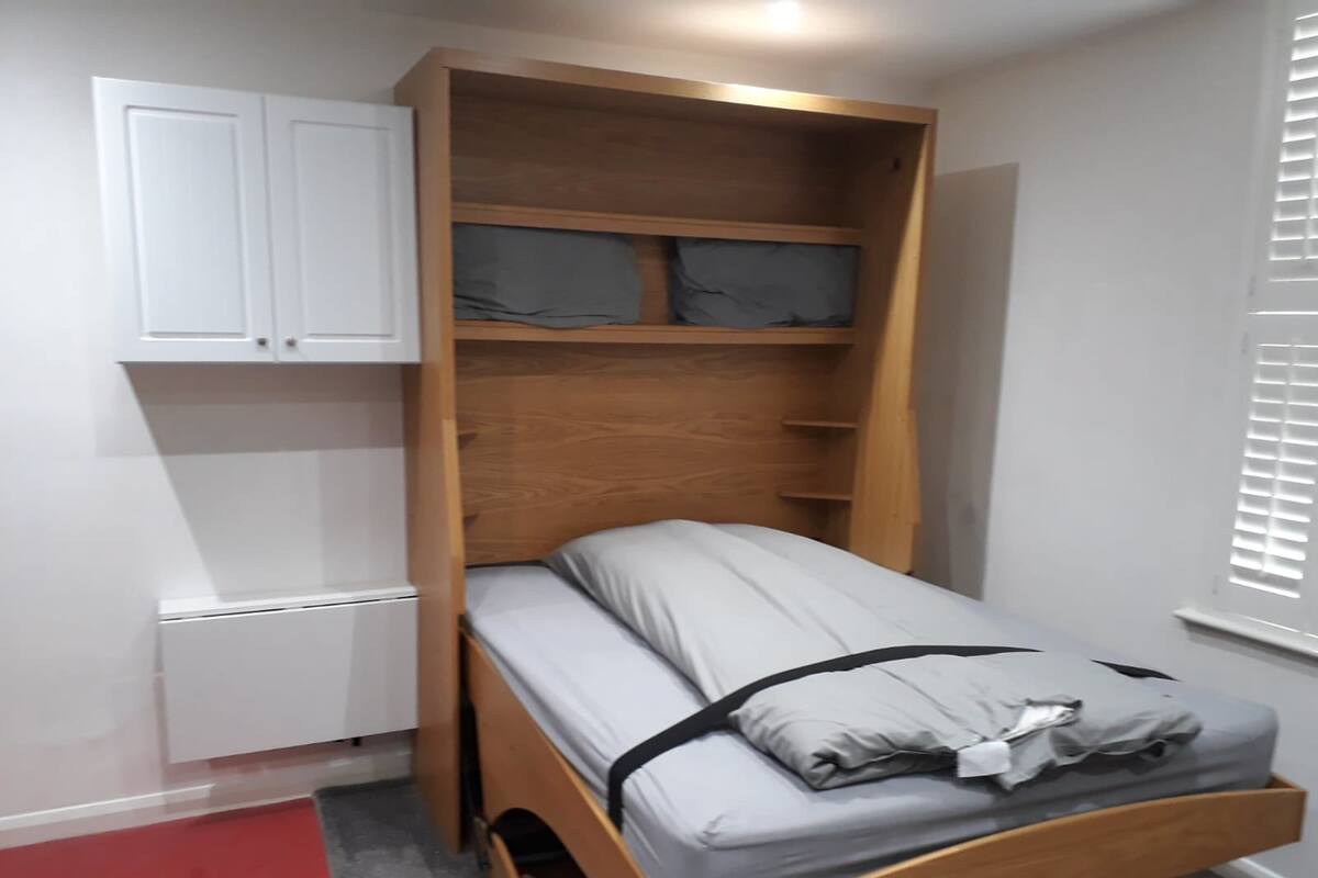 Ein kleines Zimmer mit einem Klappbett, einem kleinen Schrank und einem Fenster mit Jalousien.