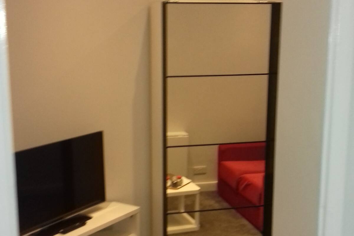 Ein Schlafzimmer mit einem großen Spiegel, einem Fernseher auf einer weißen Kommode, einem roten Bett und einem kleinen weißen Tisch.