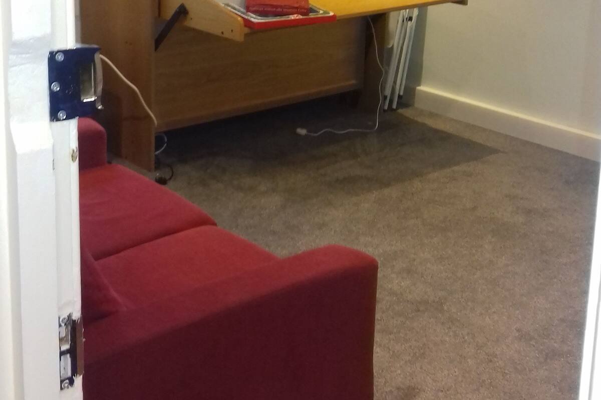 Ein kompakter Raum mit einem Klappbett, einem kleinen Schreibtisch mit einem Laptop und einem roten Sessel.